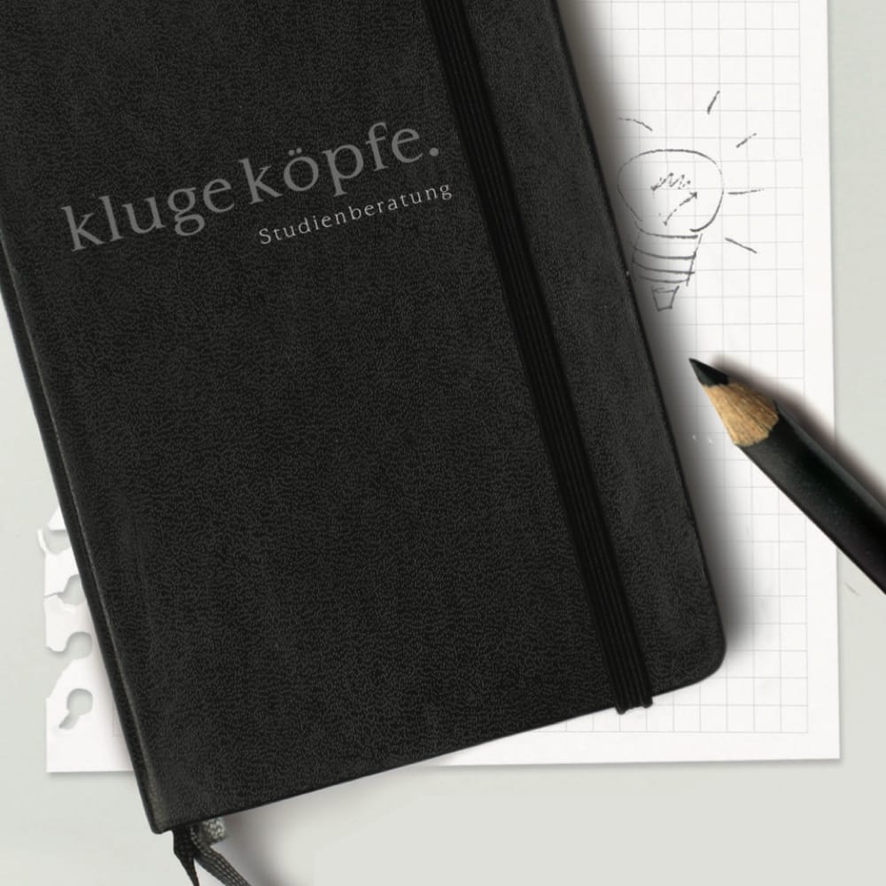 Notizbuch mit klugeköpfe Logo liegt auf kariertem Papier, daneben ein Stift